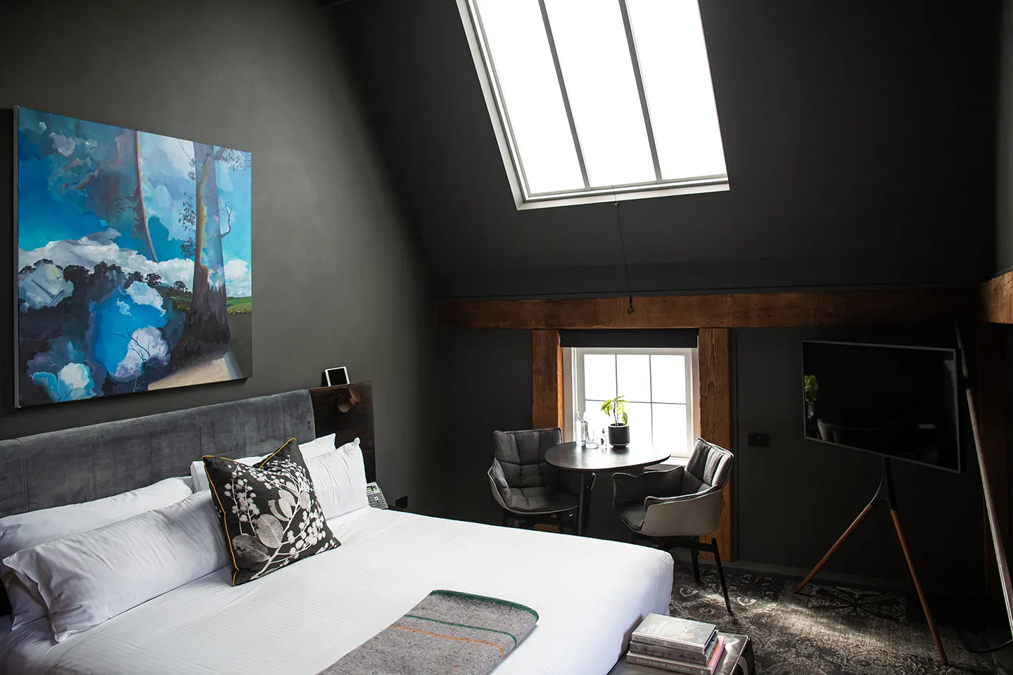 Corner of a hotel room. Skylight and window in view. In the foreground is a comfy bed, colourful art and two chairs with a table, and a elegant tv.