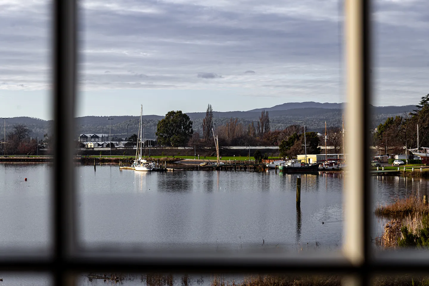 A water view through a wooden window featuring boats, water and reeds. Hills are in the background. 