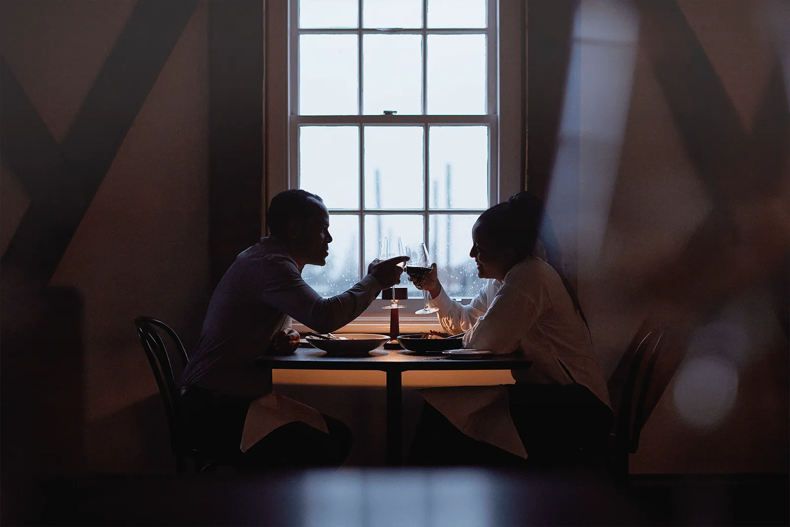 A couple sits in front of a window overlooking the river. The lighting is romantic and moody, and they are celebrating a special occasion with wine. 