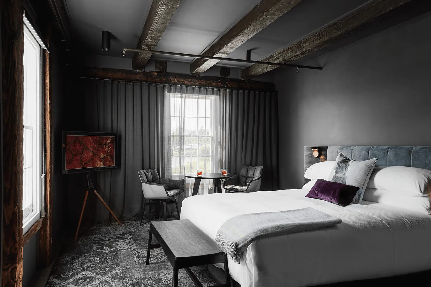Room with a bed and pillows, a desk with chairs, and a window. Walls have wooden beams and a hanging painting. Floor appears upside down.