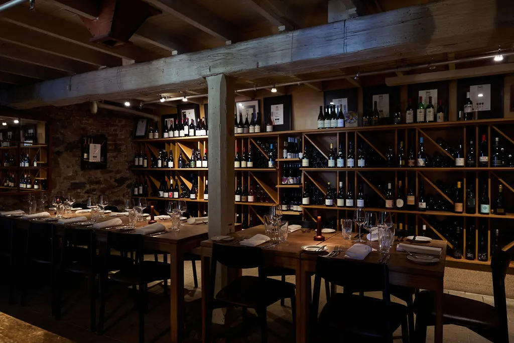 A wine cellar, with a wall of wine in the background, wooden beams and a beautifully set long table in the centre. 