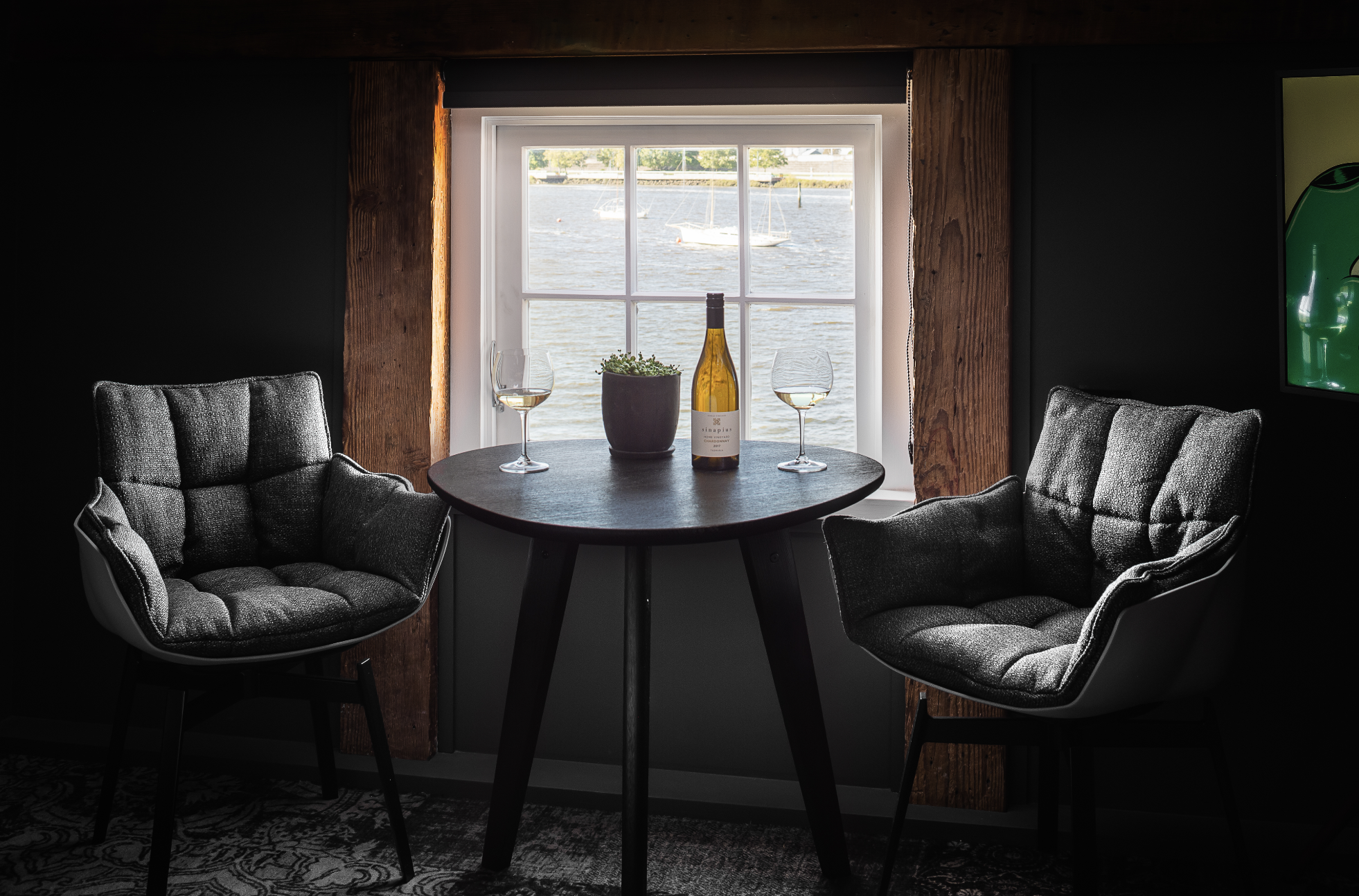 A restaurant, featuring wooden tables and chairs. Windows overlook the river. A full table setting awaits in preparation for guests. 