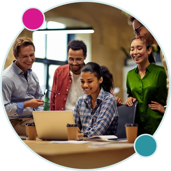 A group of five diverse colleagues gathered around a laptop at a table, smiling and engaging collaboratively in a modern office setting.
