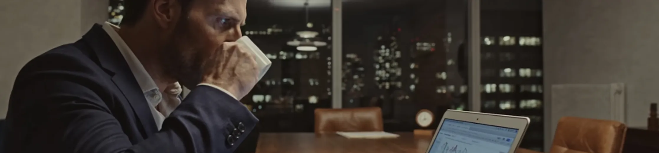 Man in a suit drinking coffee while looking at a laptop screen with charts and graphs in a dimly lit office at night with city lights visible through the windows.