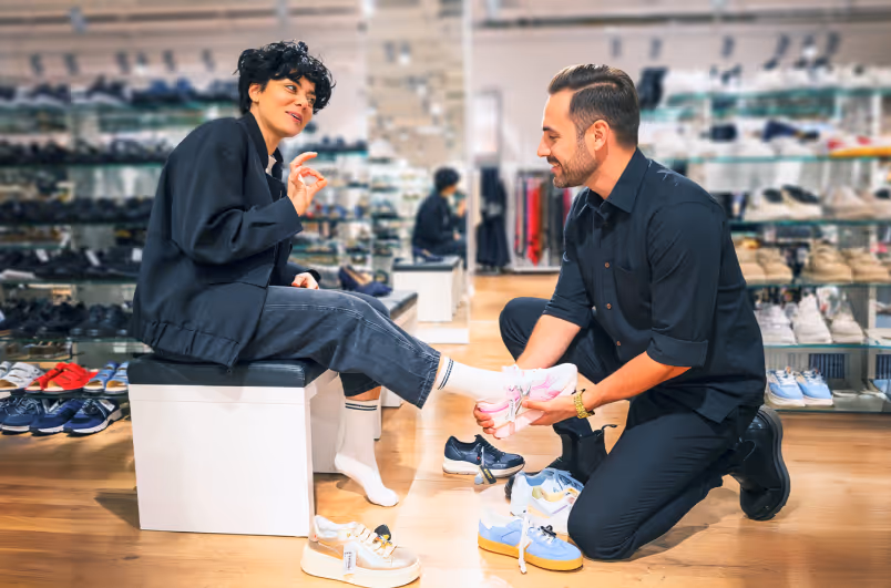 A man assists a woman trying on shoes while she sits on a bench in a shoe store.