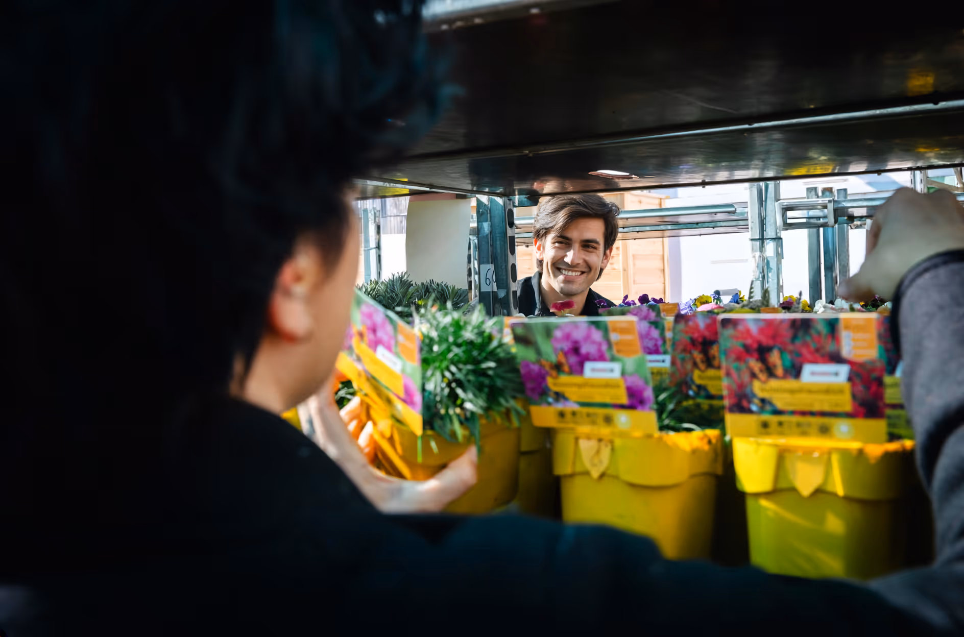 A man looks at a flower in a pot, glancing at a woman through a flower-filled alley in a shop.