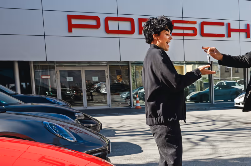 In front of a Porsche dealership, a man gives a car key to a woman.