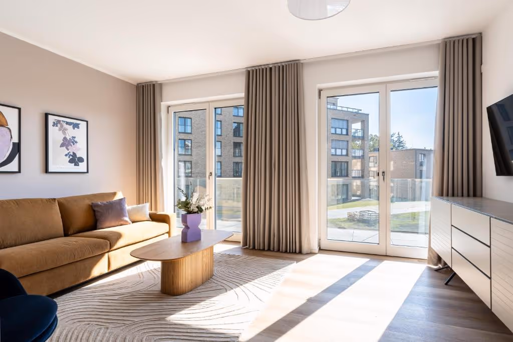 Helles, modernes Wohnzimmer mit braunem Sofa, Holz-Couchtisch, großen Fenstern mit Blick auf Außengelände und Vorhängen.