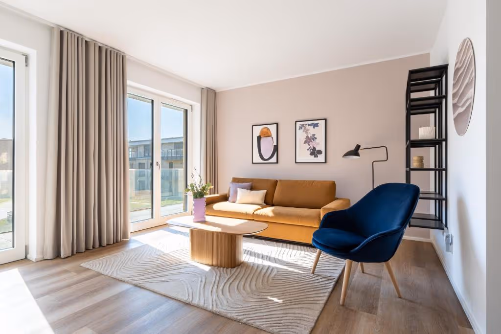 Helles Wohnzimmer mit orangefarbenem Sofa, blauer Sessel, modernem Couchtisch und großen Fenstern mit Vorhängen.