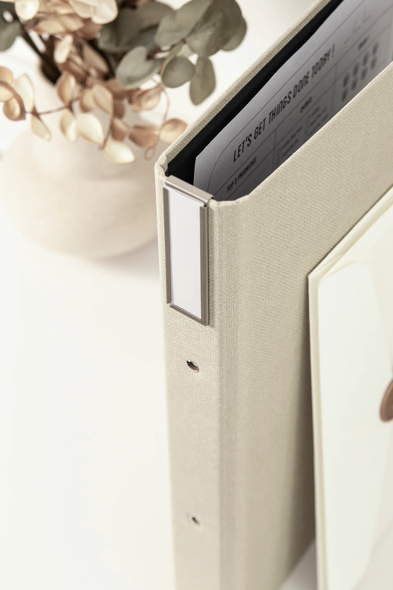 Beige fabric-covered binder from Bigso featuring a sleek detachable label holder clip on the spine, designed for easy organization and customization. A vase with dried leaves decorates the background, highlighting the stylish and practical storage solution from Bigso’s News article on label holder clips.