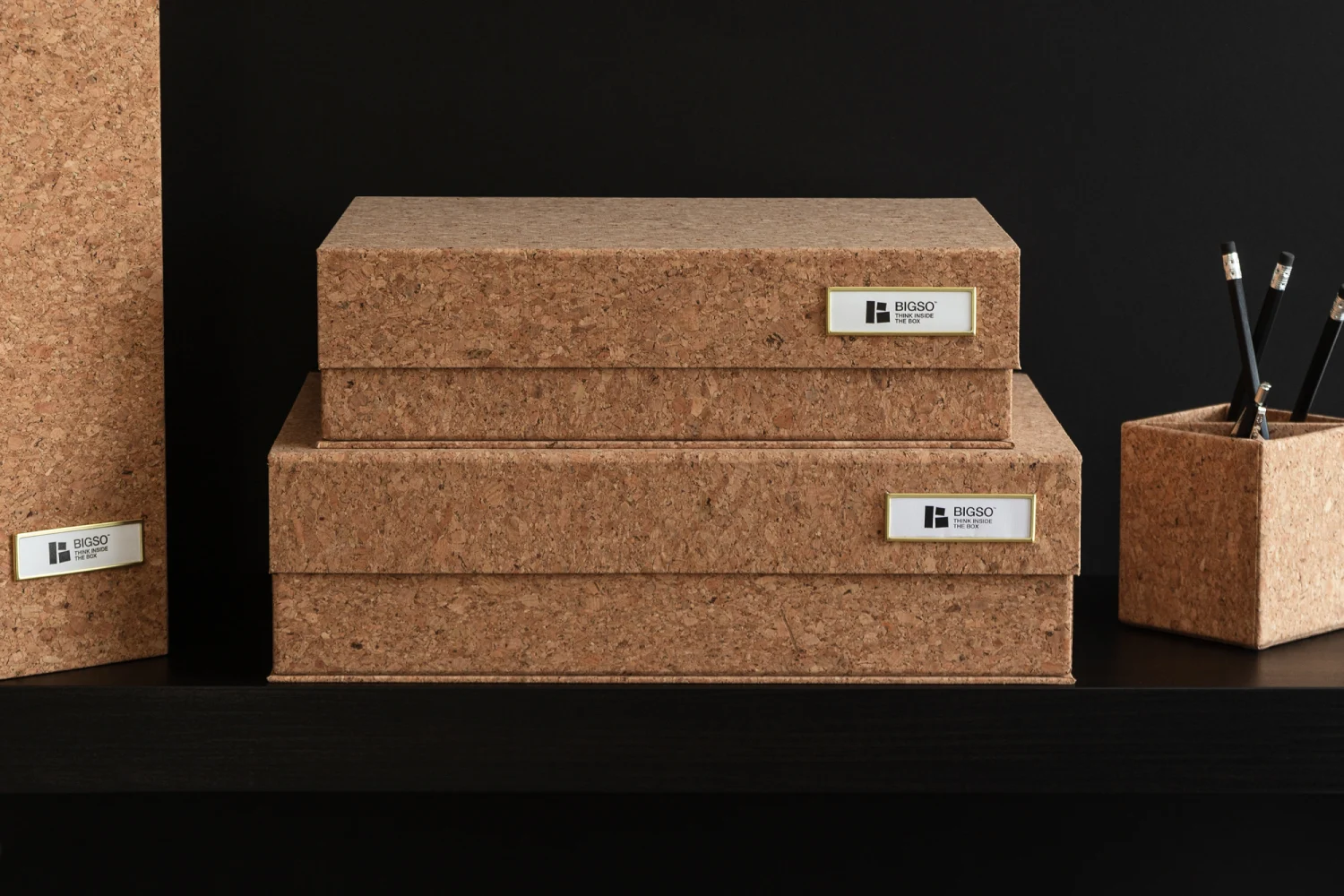 Bigso cork storage boxes and magazine file with label holders, displayed on a black shelf. Eco-friendly Scandinavian storage solution made from natural cork and recycled fiberboard, combining sustainable materials with modern minimalist design for home and office organization.