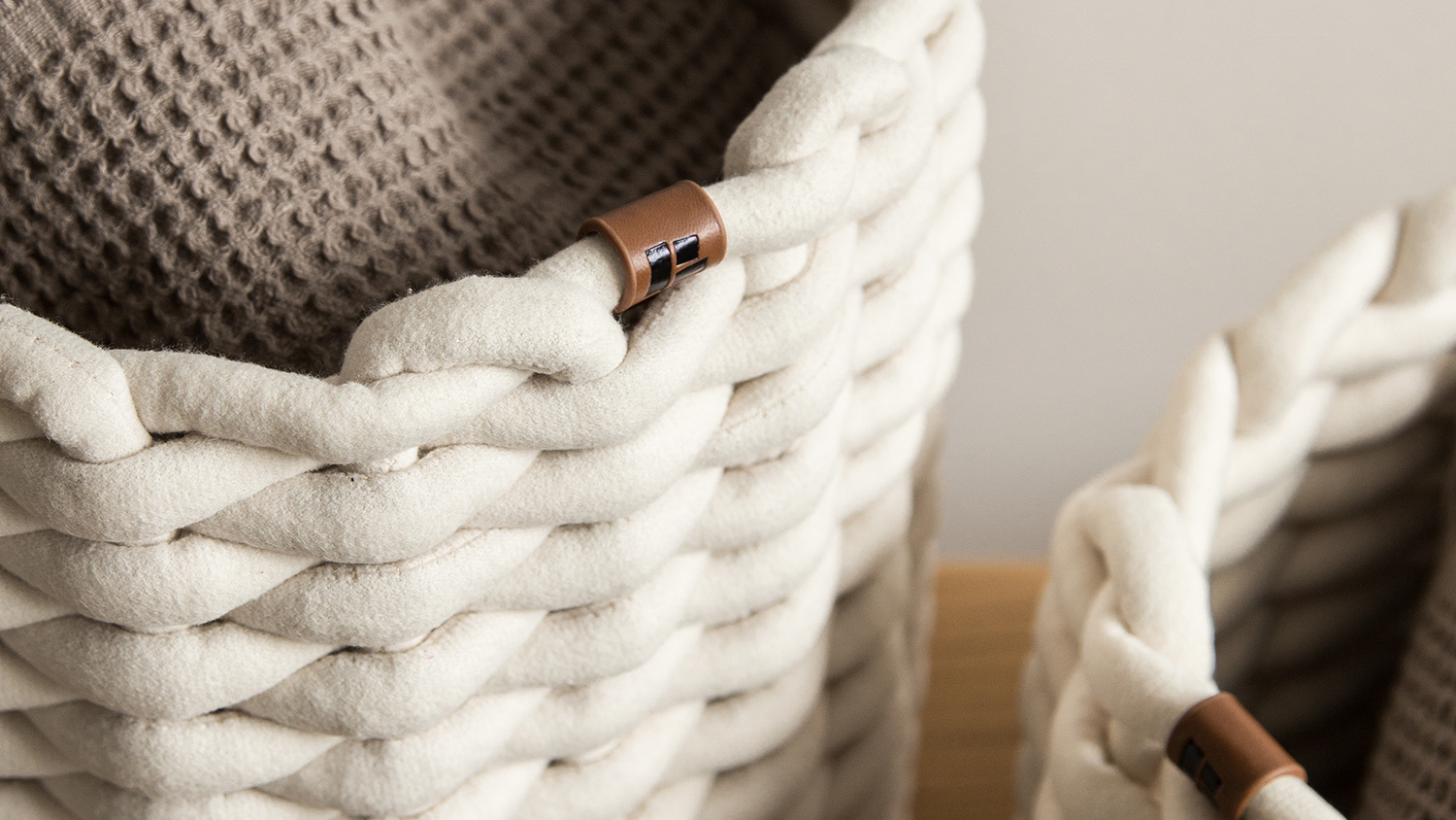 Grey braided storage basket made from peach skin fabric with padded rope design and brown leather tag, styled with blanket in modern home interior