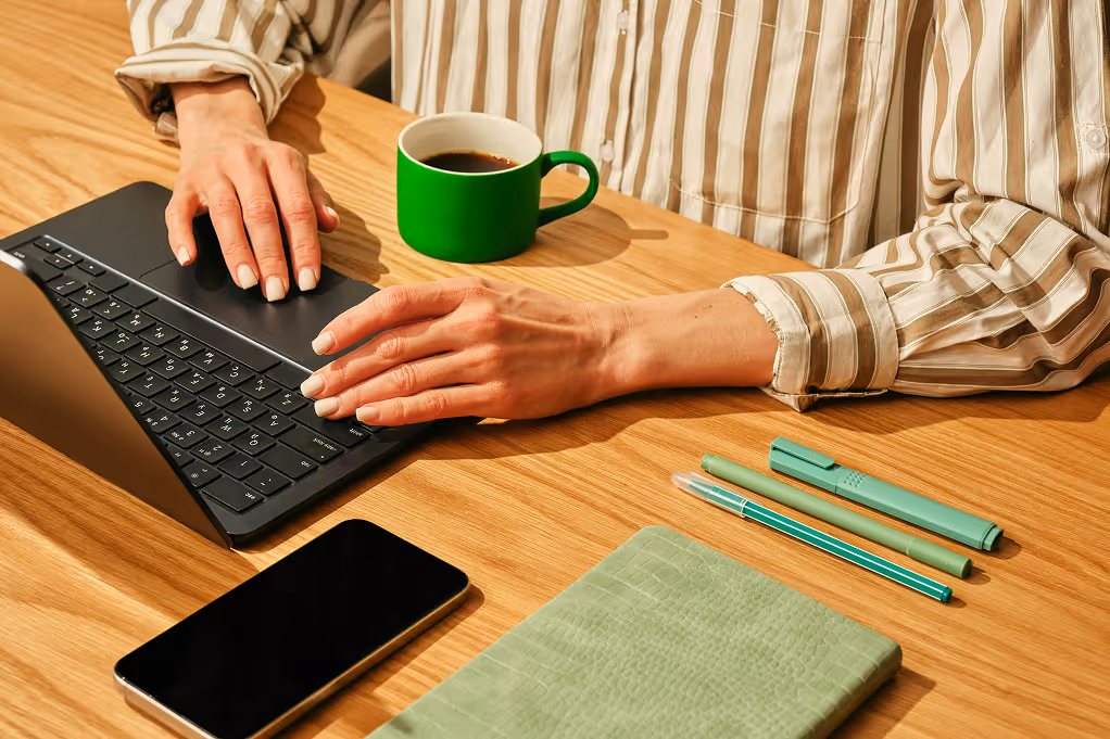 Mains d'une personne tapant sur un clavier d'ordinateur portable noir avec une tasse verte, un smartphone et des stylos sur une table en bois.