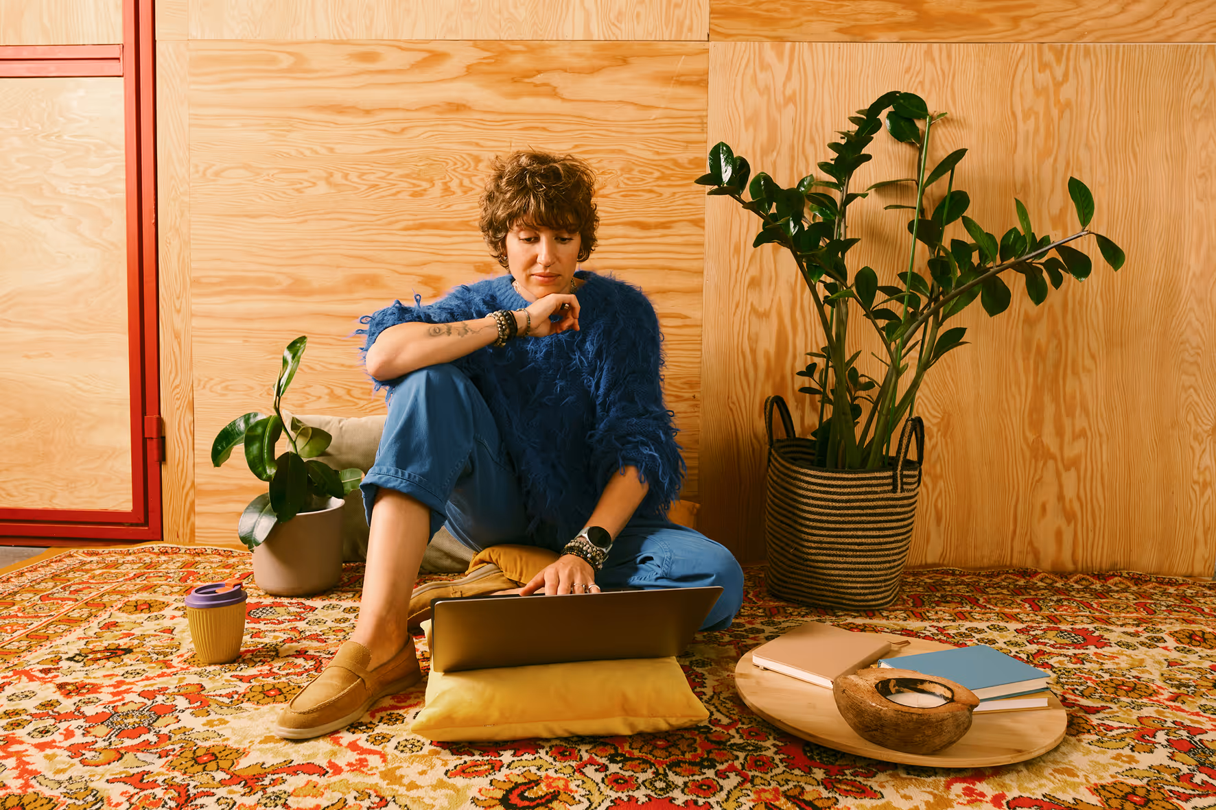 Personne assise par terre sur un tapis coloré avec un ordinateur portable, entourée de plantes d'intérieur et d'objets décoratifs.