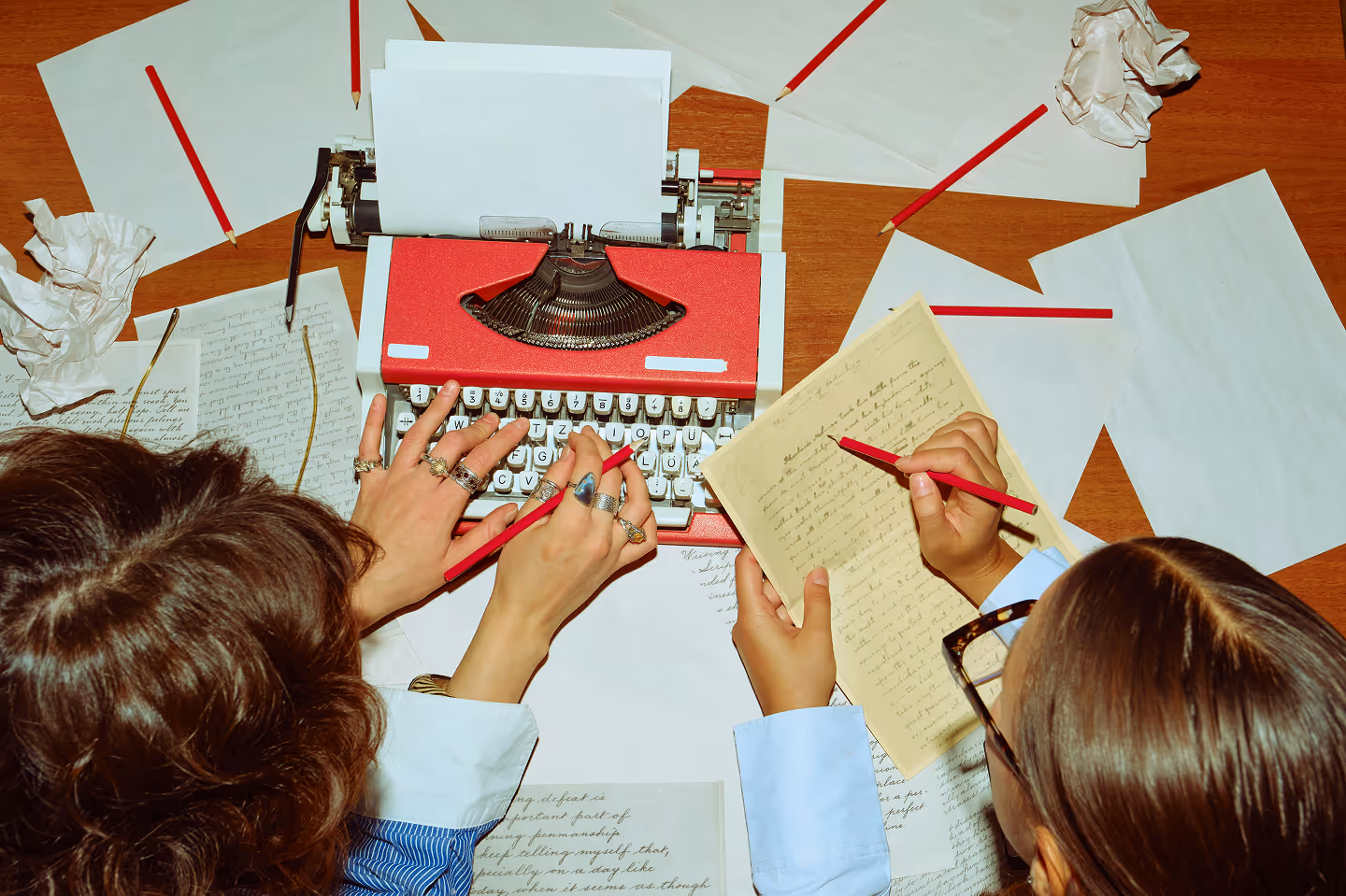 Deux personnes tapant à la machine à écrire rouge avec des papiers et des crayons rouges éparpillés sur une table en bois.