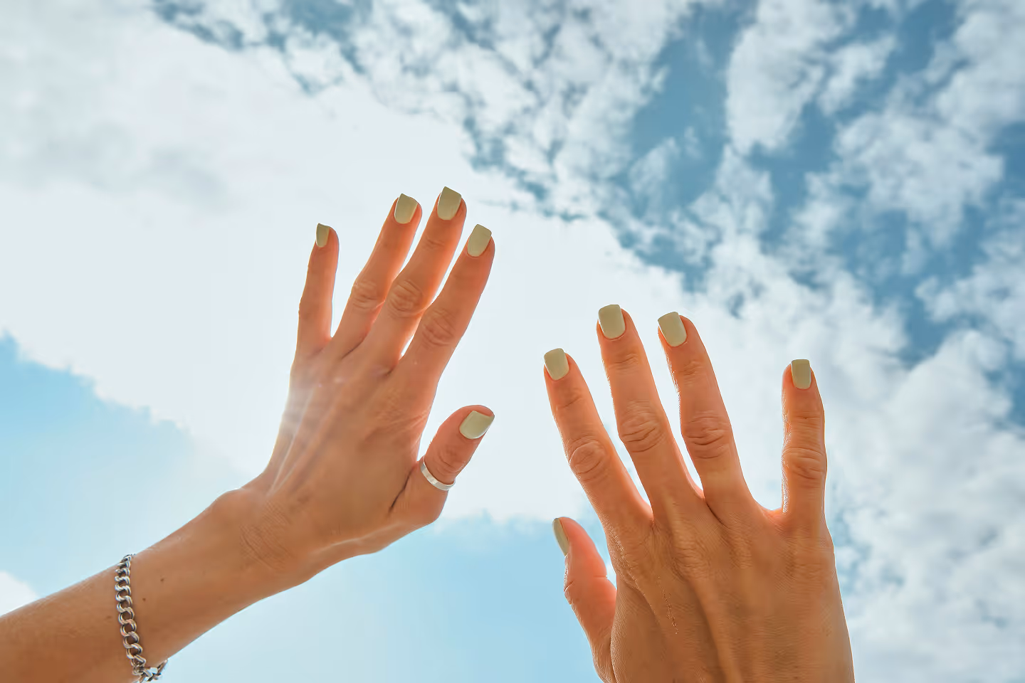 Mains féminines levées vers un ciel partiellement nuageux avec des ongles peints en vert clair.