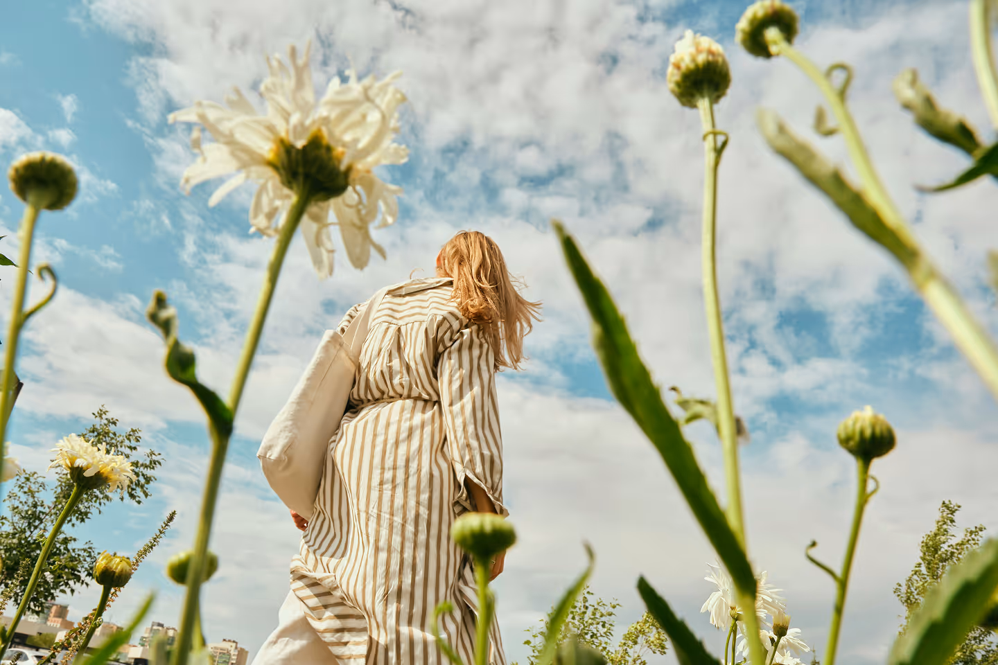 Femme vêtue d'une robe rayée tenant un sac beige, vue en contre-plongée parmi des grandes fleurs blanches sous un ciel partiellement nuageux.