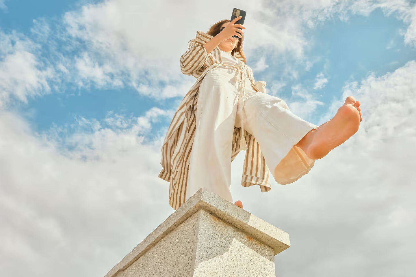 Femme debout pieds nus sur une base en pierre, prenant une photo avec un smartphone sous un ciel bleu partiellement nuageux.