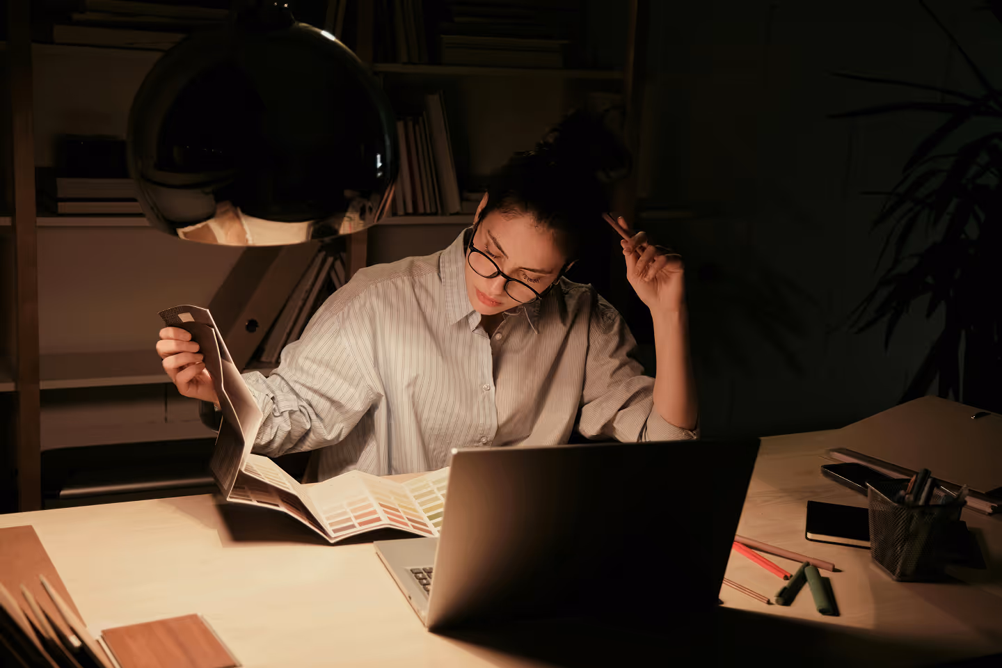 Jeune femme portant des lunettes, travaillant tard le soir avec des échantillons de couleurs et un ordinateur portable sous une lampe de bureau.