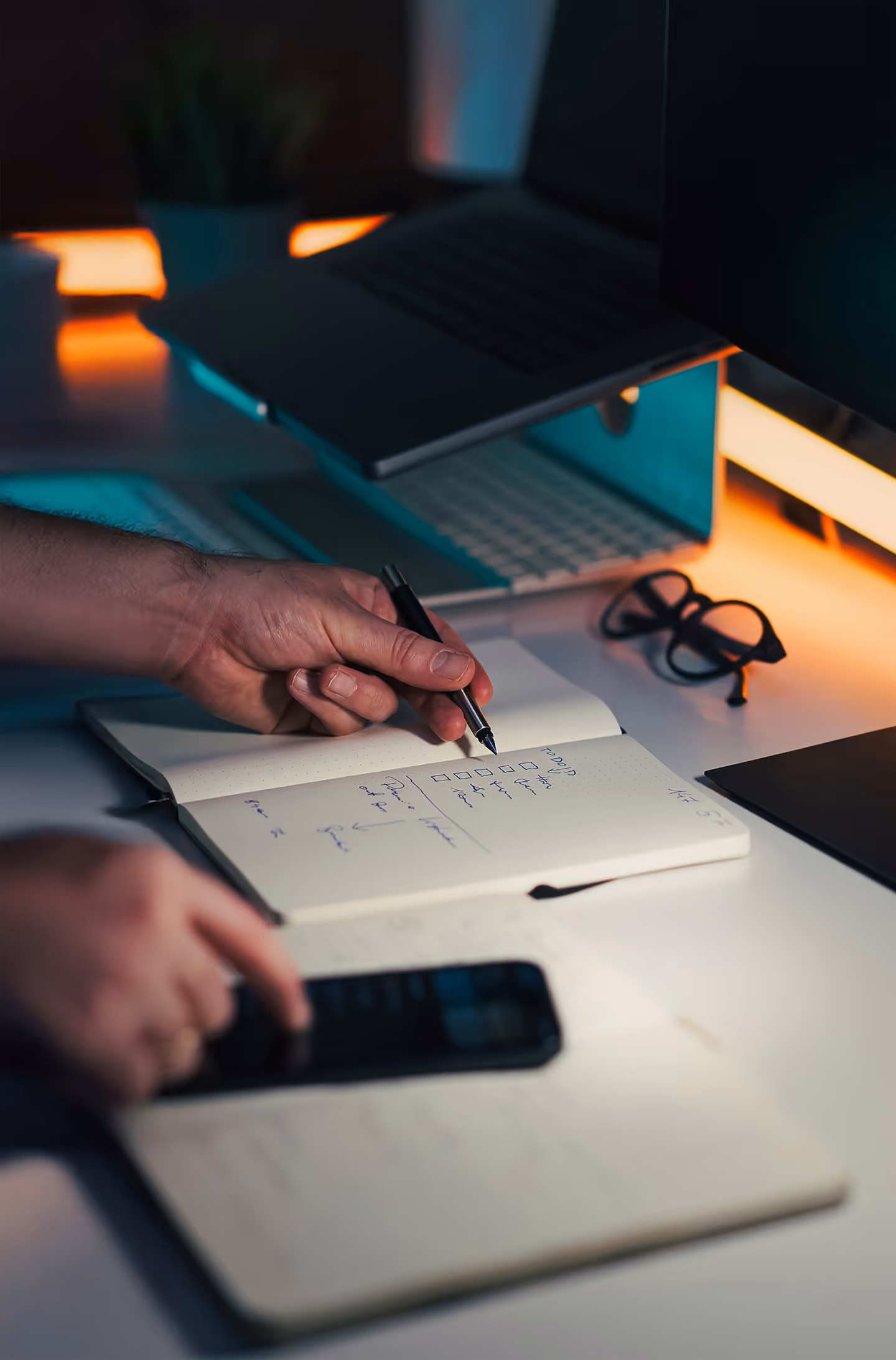 Personne notant une liste de tâches dans un carnet avec un ordinateur portable et des lunettes sur un bureau éclairé.