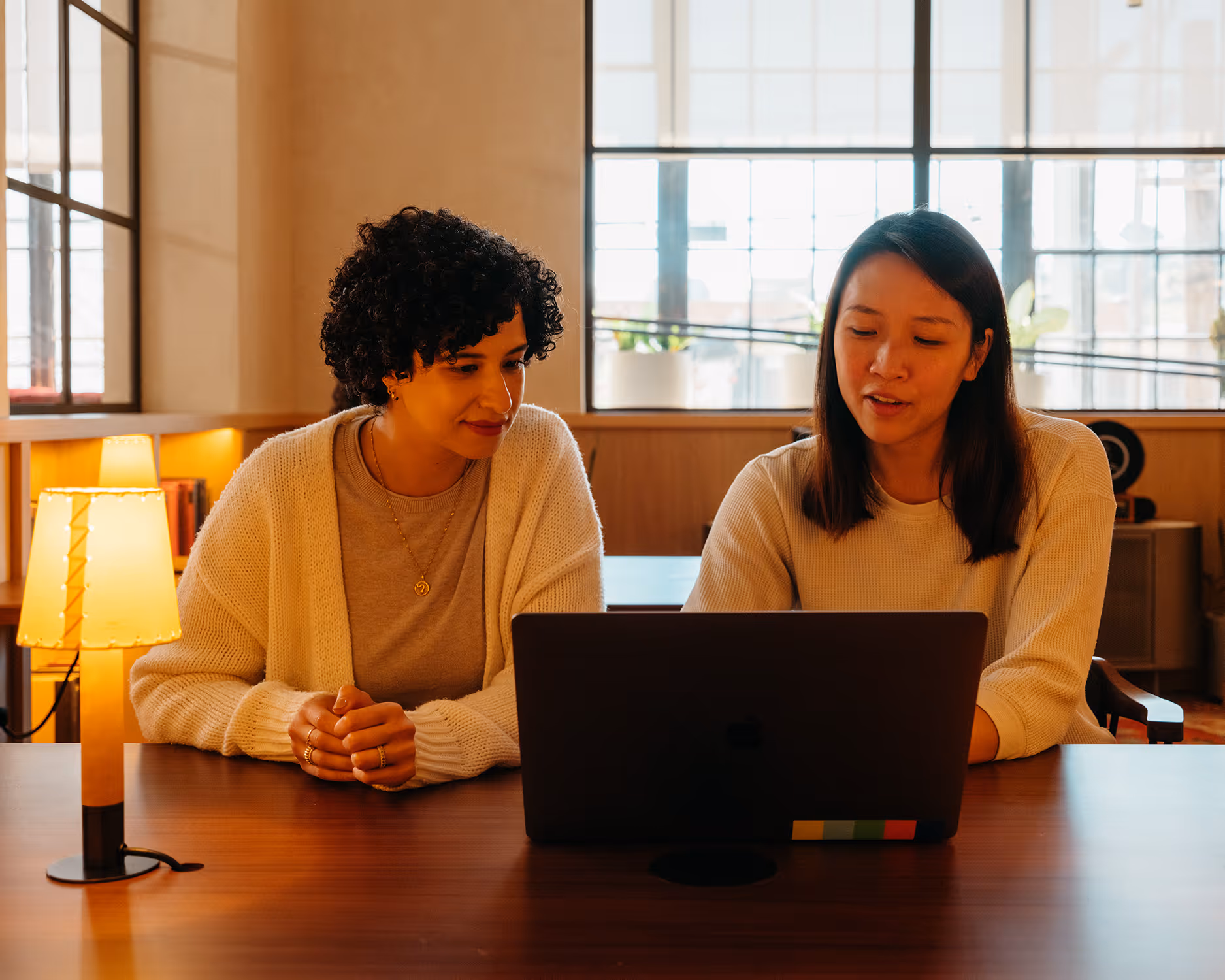 Deux femmes collaborant ensemble autour d'un ordinateur portable dans un bureau lumineux avec une lampe sur la table.