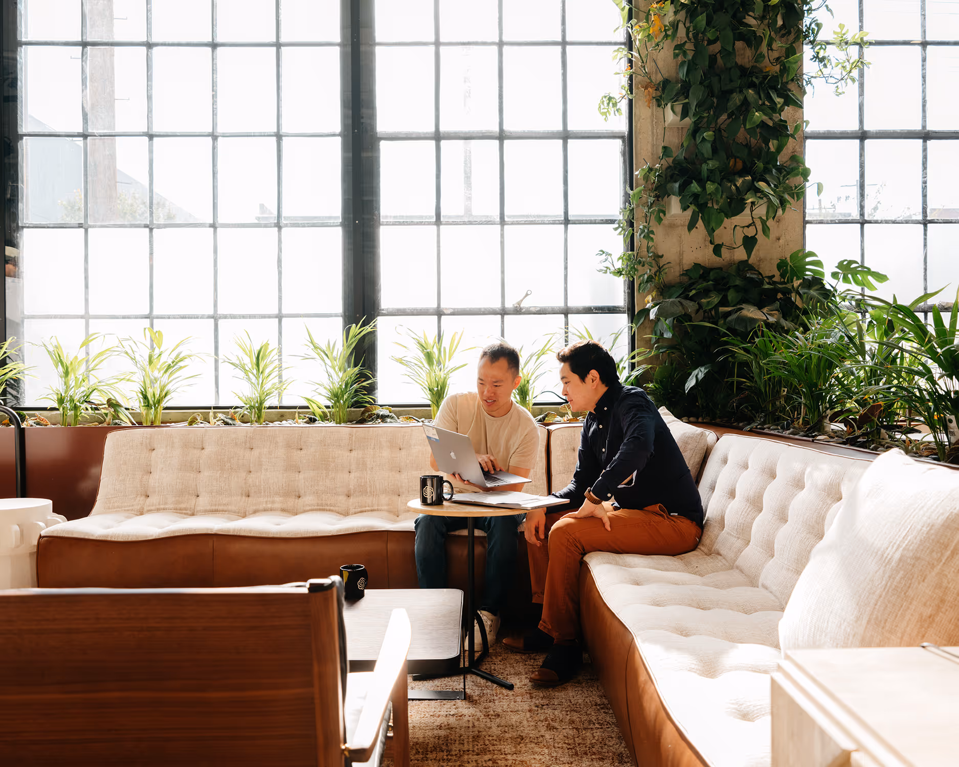 Deux hommes assis sur un canapé dans un salon lumineux avec des plantes, regardant un ordinateur portable.