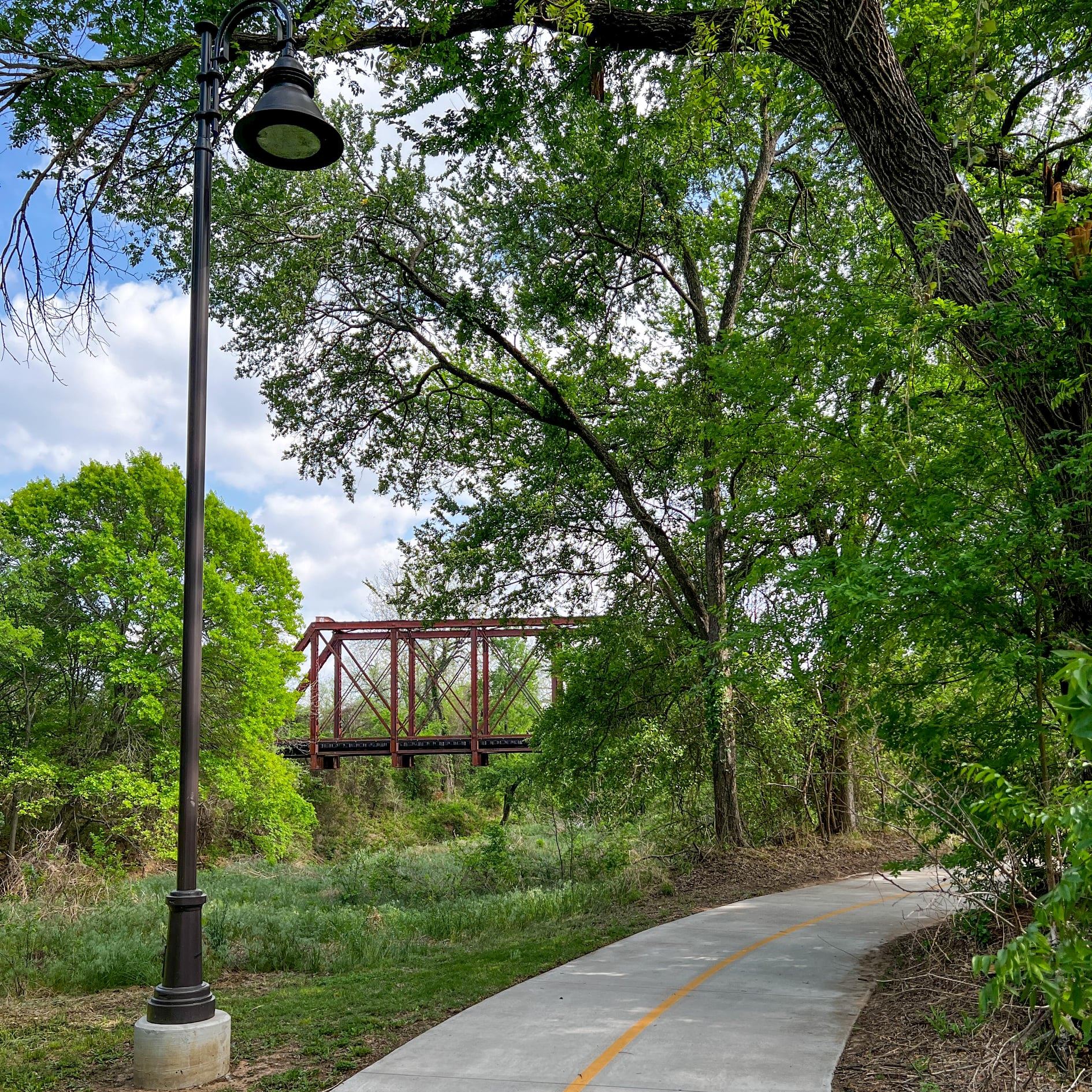 Curving paved trail lined with green trees and a black street lamp, with a rusted red metal bridge in the background.