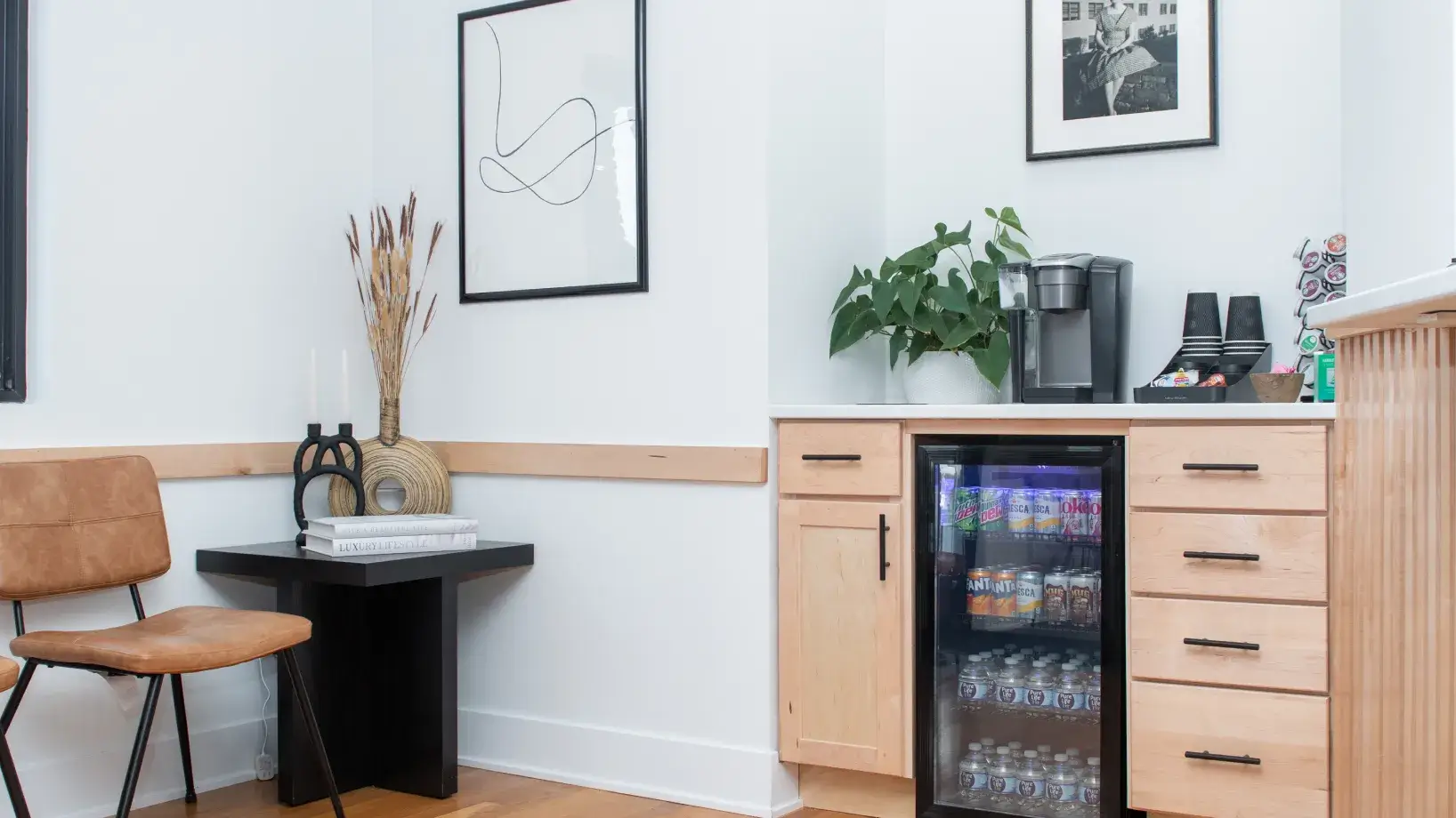 Modern kitchen corner with wooden cabinets, a mini fridge stocked with drinks, a coffee maker, and a seating area with a tan chair and black side table decorated with books and vases.