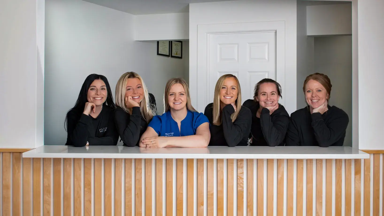 Six women smiling and leaning on a reception desk with wood paneling and white countertop in a bright office.