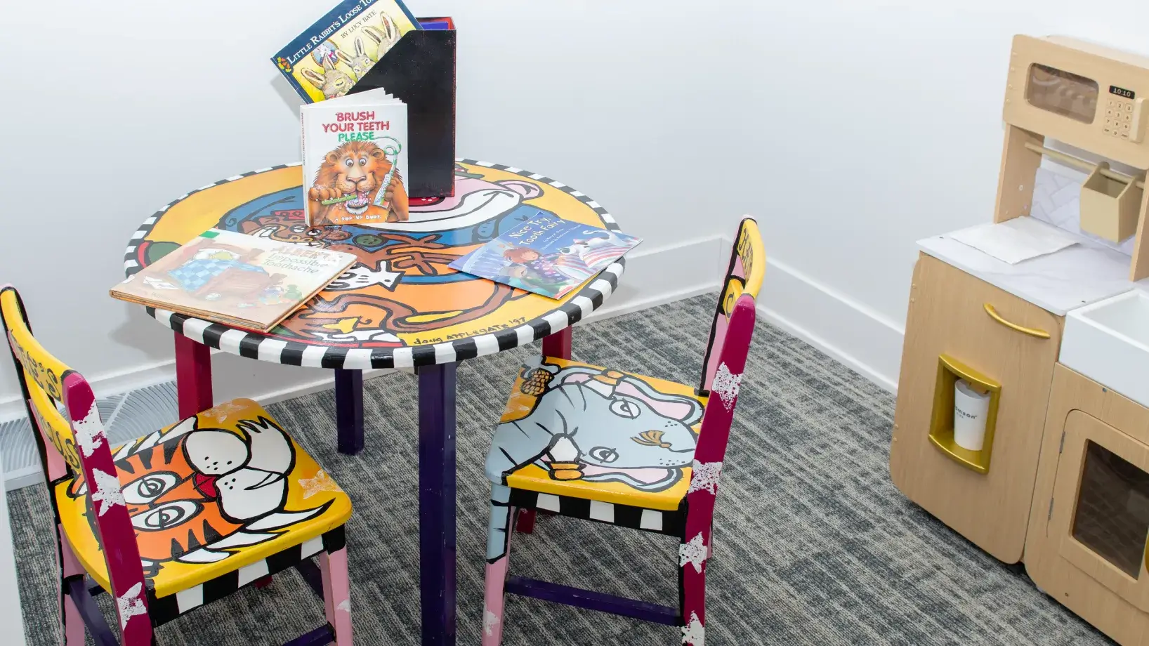 Children's play area with a colorful round table and two painted animal chairs, alongside a wooden play kitchen set.