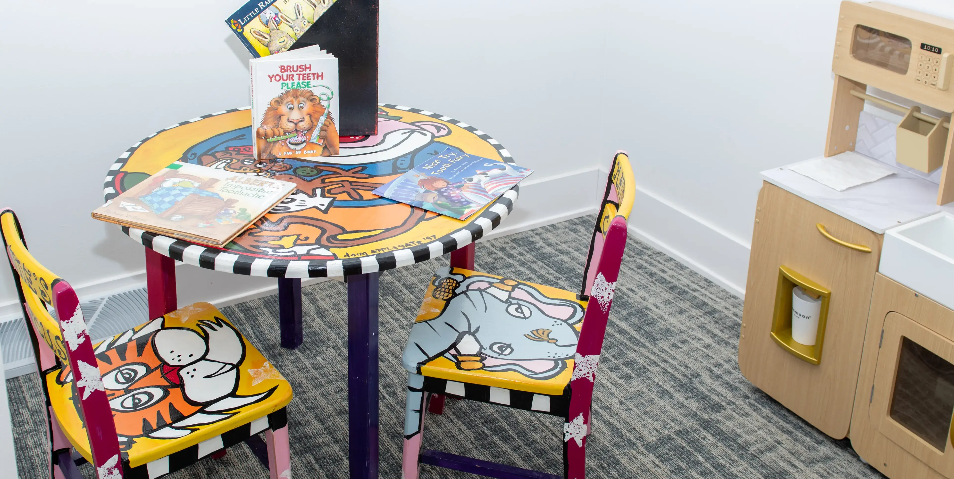 Children’s play area with a colorful round table and two chairs featuring animal designs, books on dental hygiene placed on the table, and a wooden play kitchen set in the corner.