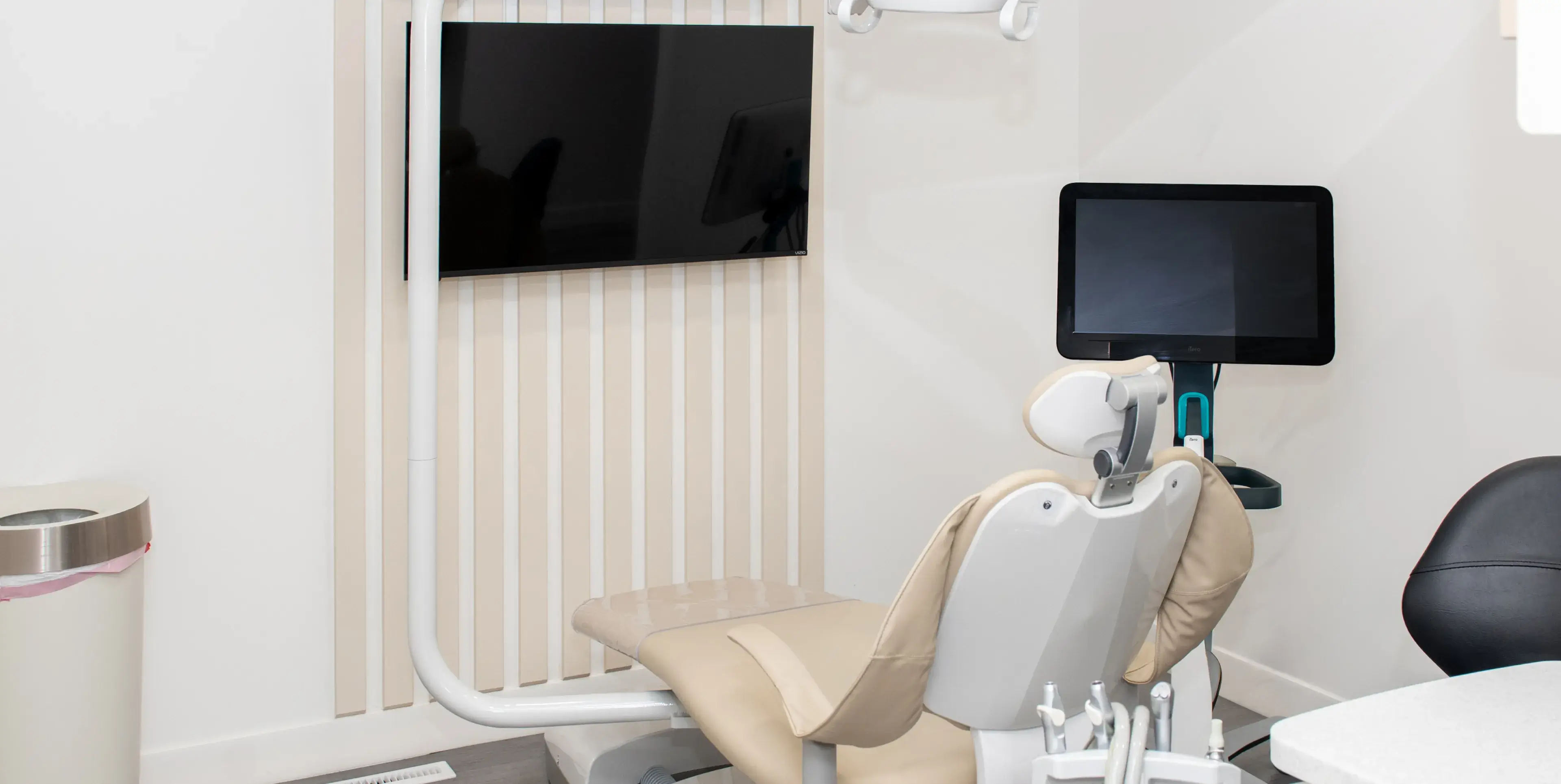 Modern dental examination room with beige dental chair, monitor screens, and dental tools.