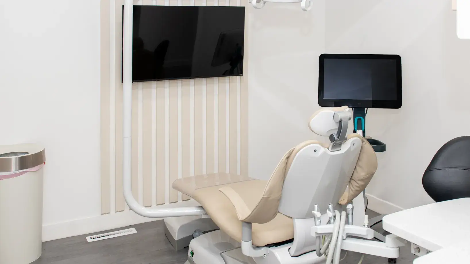 Modern dental clinic room with beige dental chair, medical equipment, and two mounted screens on white walls.