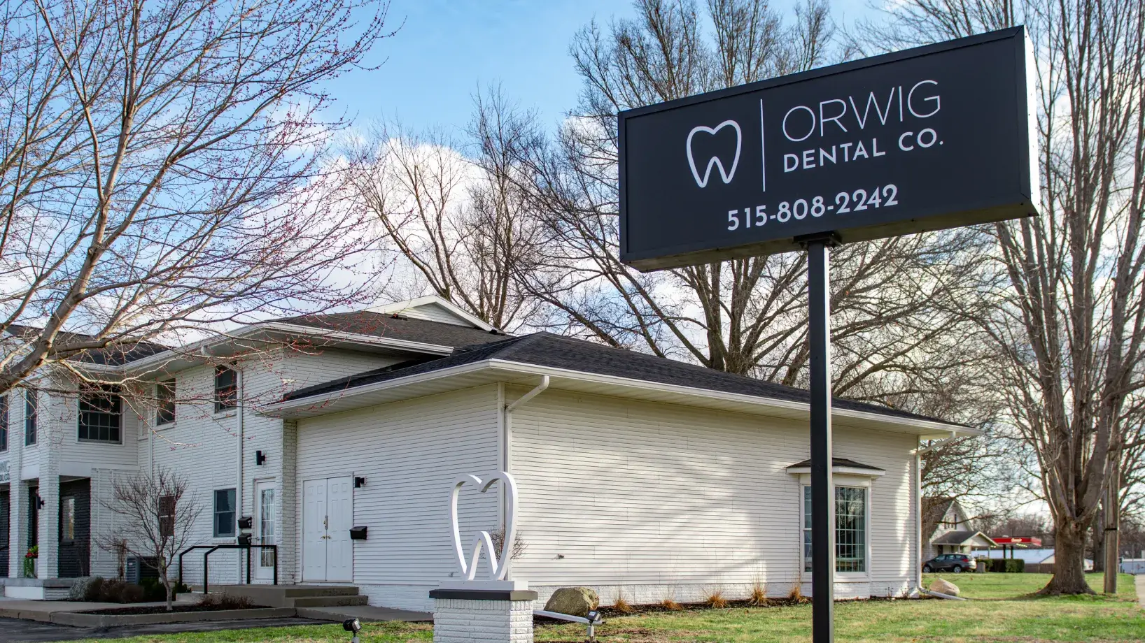 Exterior of Orwig Dental Co. building with white siding, a large black sign showing the company name and phone number, and a tooth-shaped sculpture in front.
