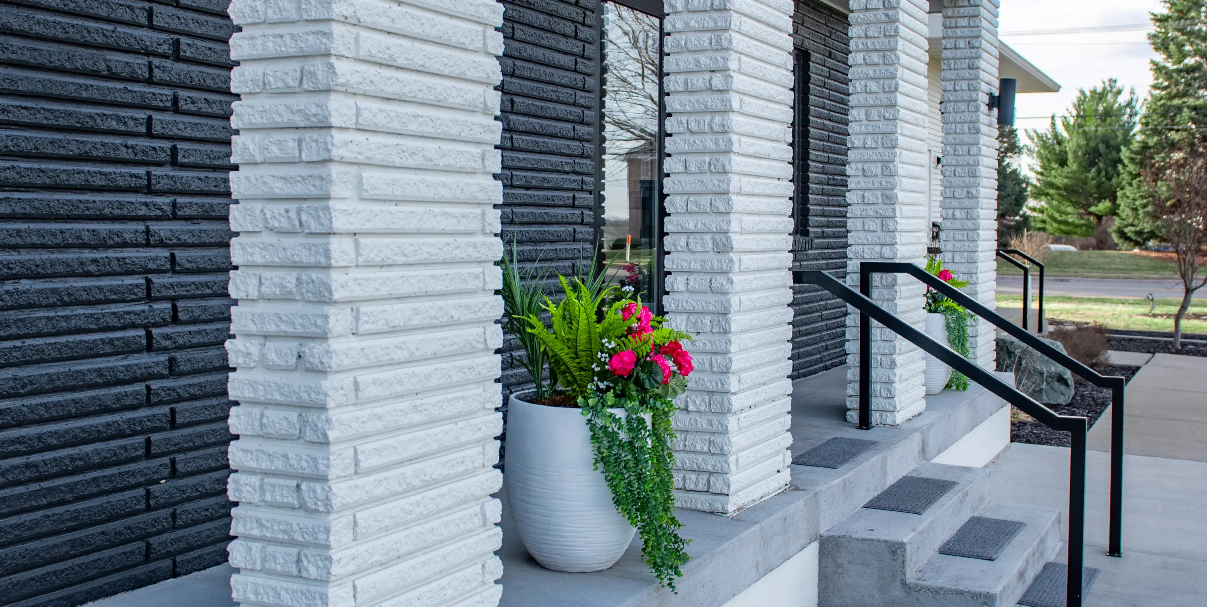 Front porch with white brick pillars, concrete stairs with black metal railings, and a large white planter with green plants and pink flowers.