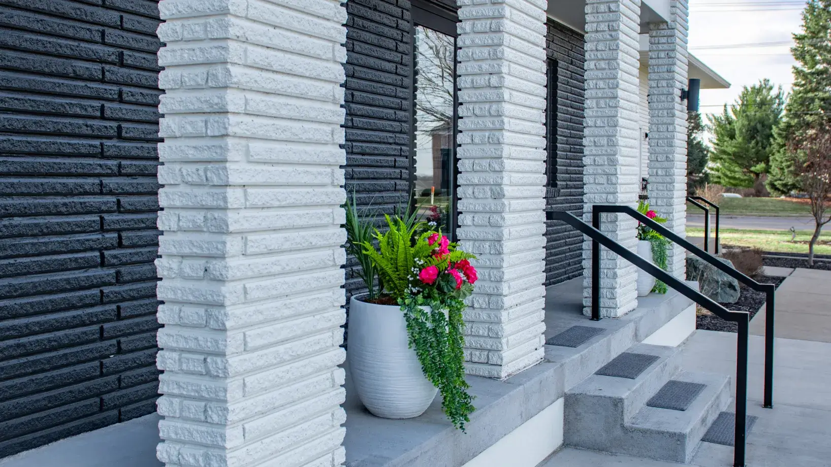 Modern house entrance with white brick columns, black metal railings, concrete steps, and large planters with green and pink flowers.