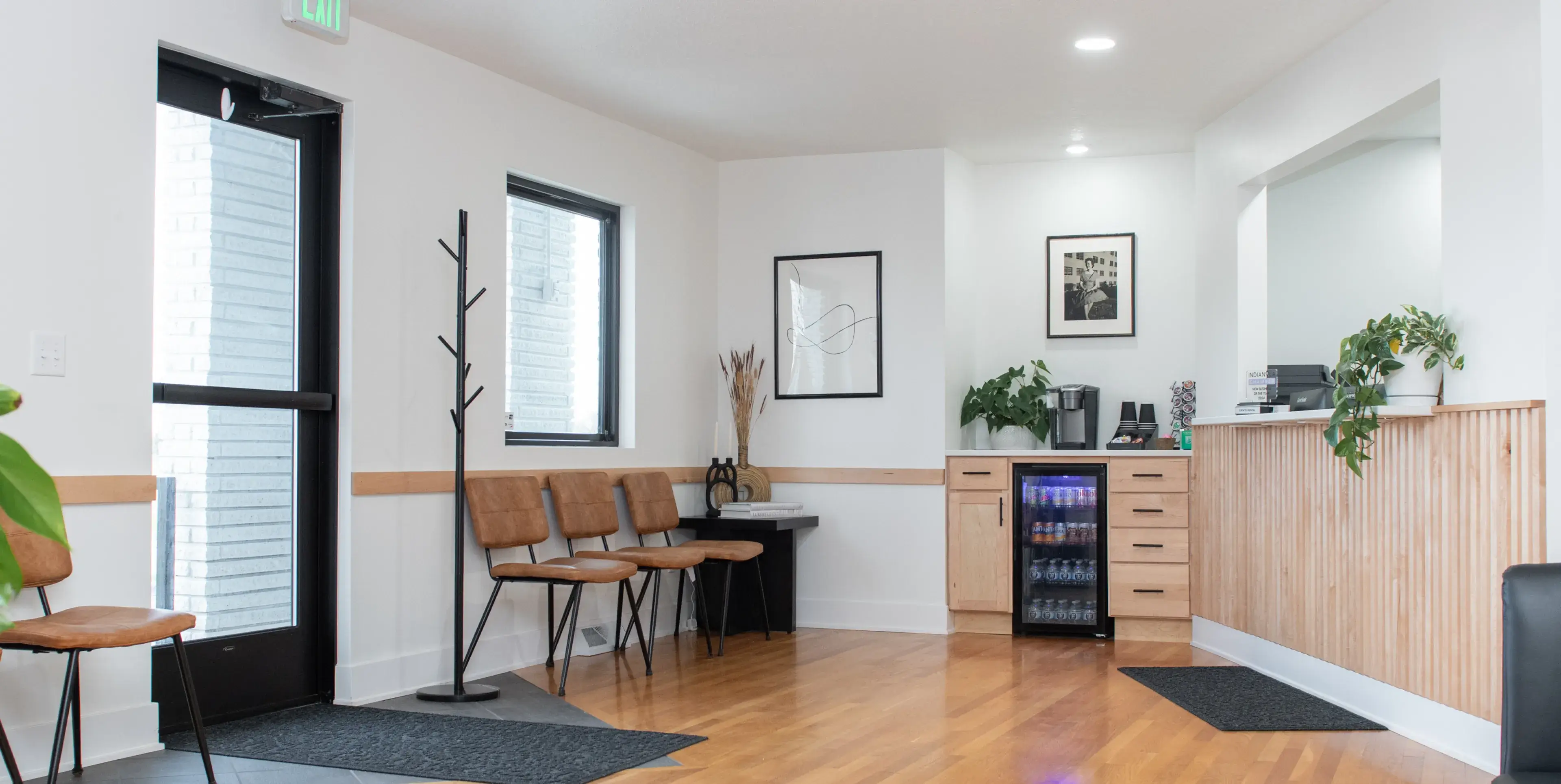 Modern waiting area with tan chairs, a coat rack, a small table, framed artwork, a beverage refrigerator, and a wooden reception counter with plants.