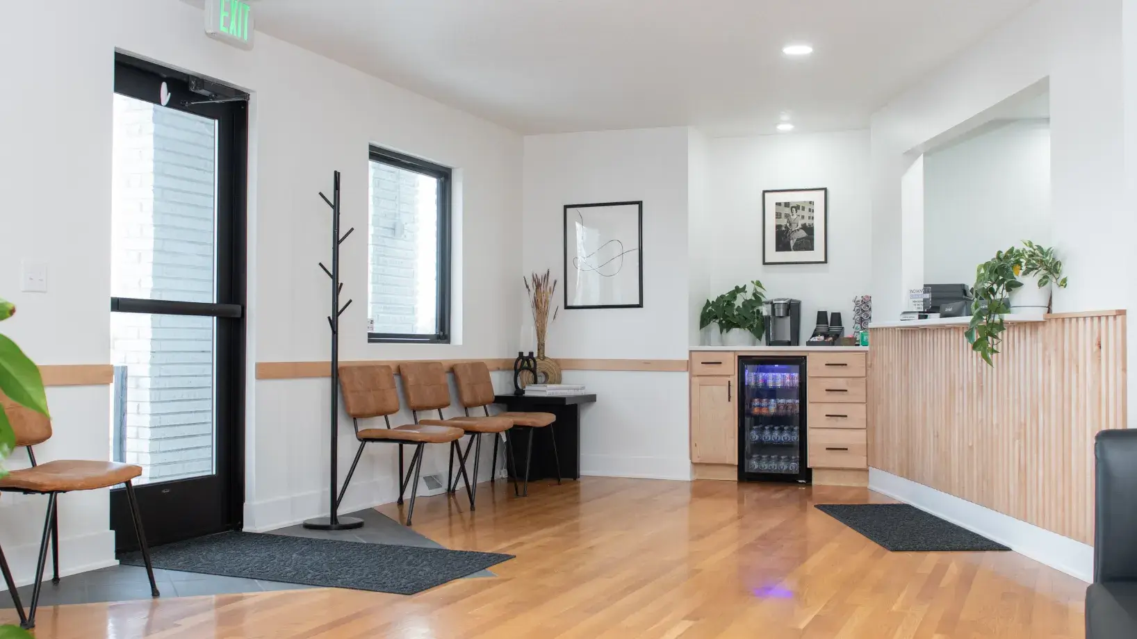 Modern, bright waiting room with wooden floor, brown chairs, coat rack, beverage fridge, coffee station, and plants.