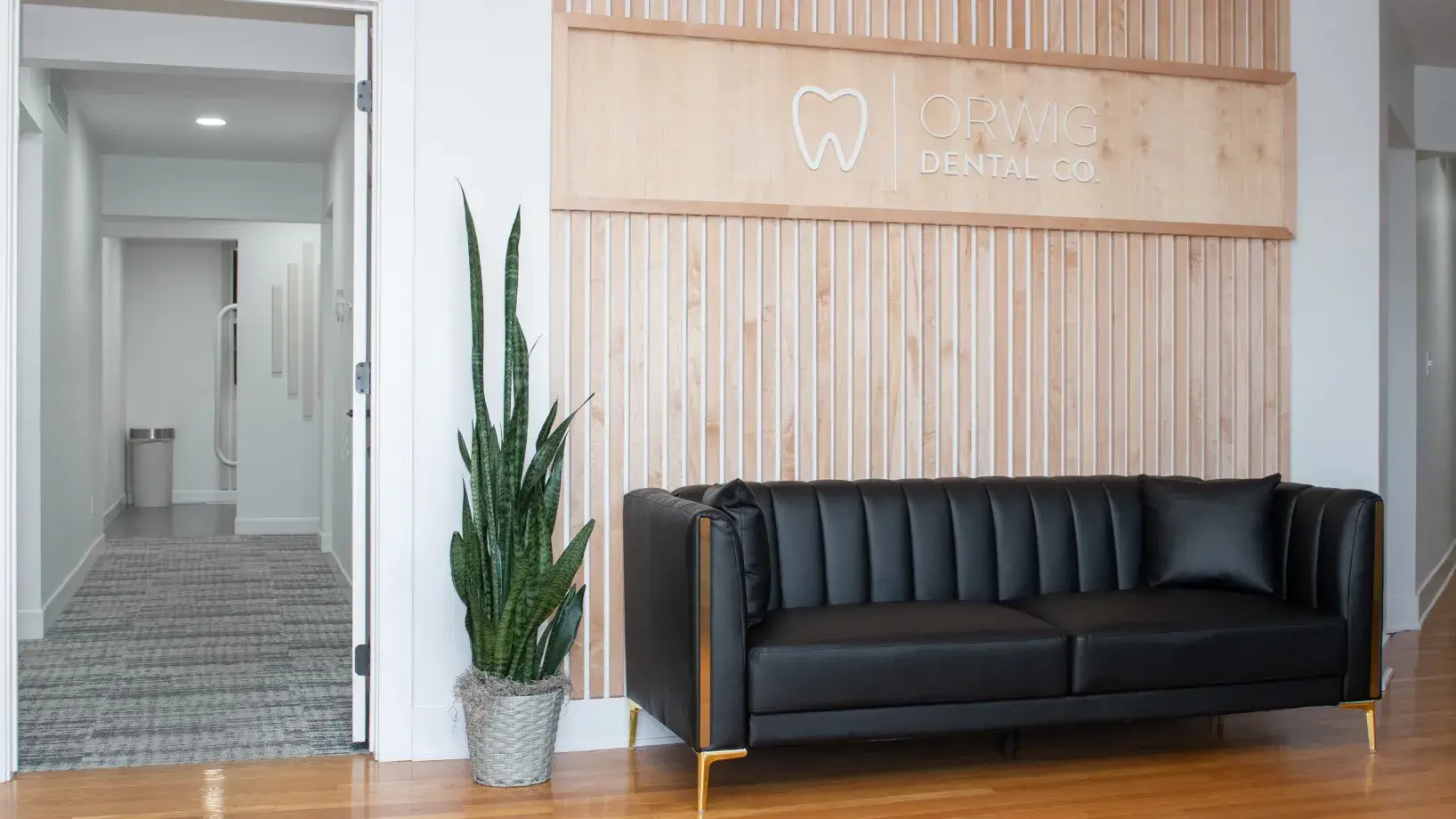 Modern dental office waiting area with a black leather couch, tall green plant, and wooden slat wall featuring Orwig Dental Co. logo.