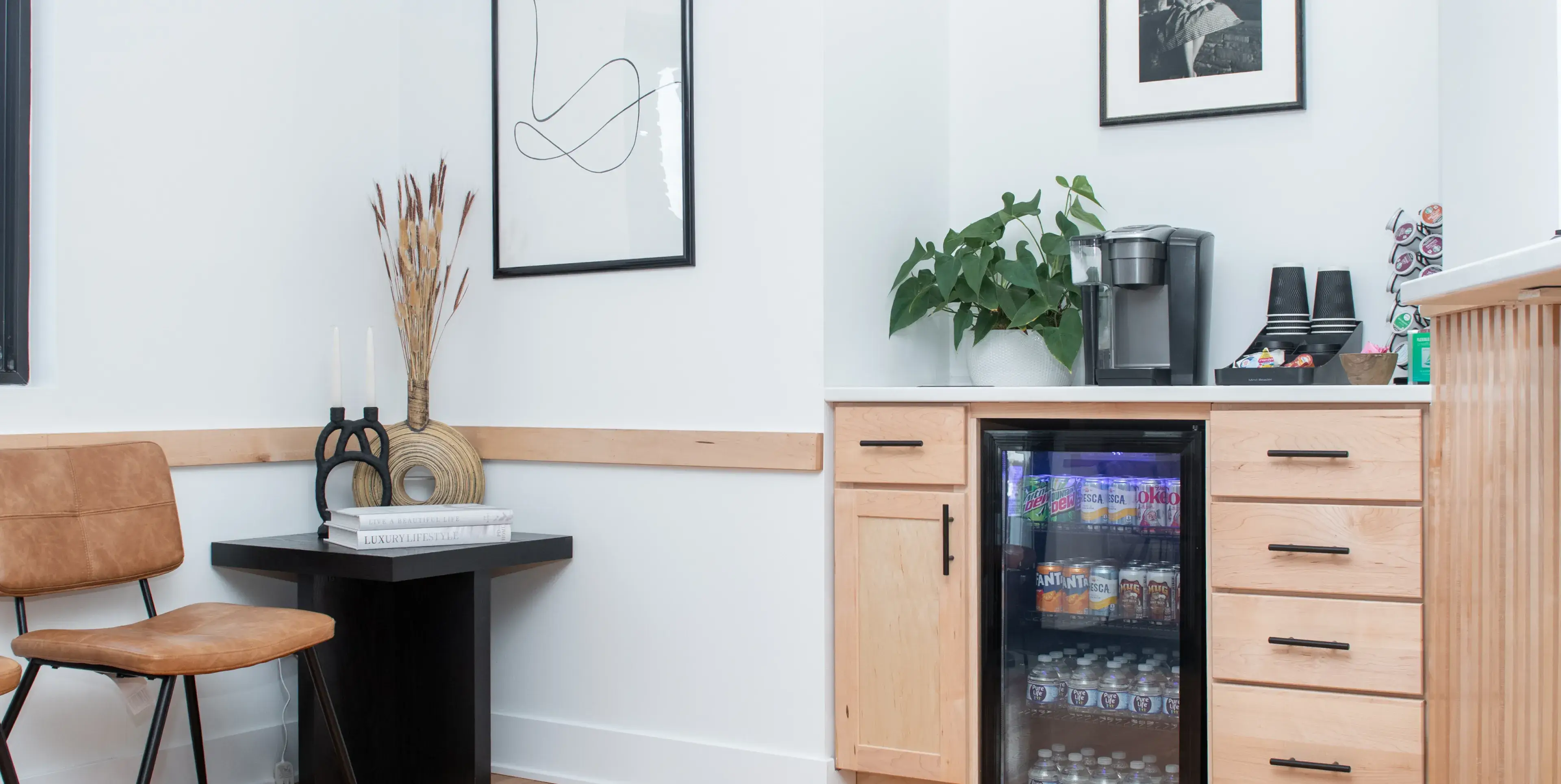 Modern office break area with a small refrigerator stocked with drinks, coffee machine, plant, and brown leather chair next to a black side table with decorative items.