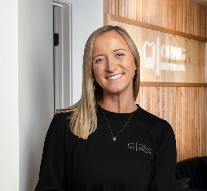 Smiling woman with straight blonde hair wearing a black ORWIG Dental Co. shirt standing indoors against a wooden wall with the ORWIG Dental Co. logo.