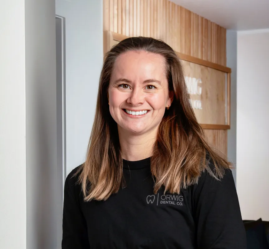 Smiling woman with long brown hair wearing a black Orwig Dental Co. shirt standing indoors.