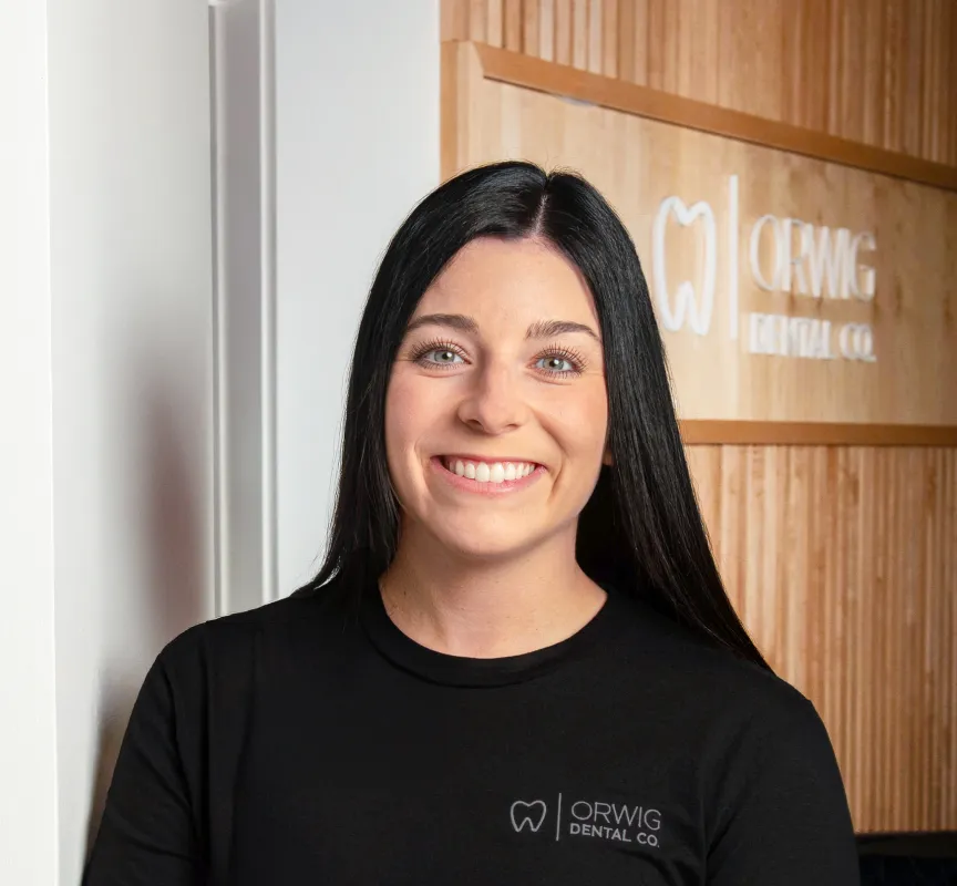 Smiling woman with long black hair wearing an Orwig Dental Co. shirt standing in front of a wooden wall with the company logo.