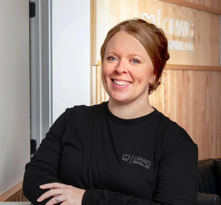 Smiling woman with red hair wearing a black Orwig Dental Co. shirt standing in an office with wooden paneling.