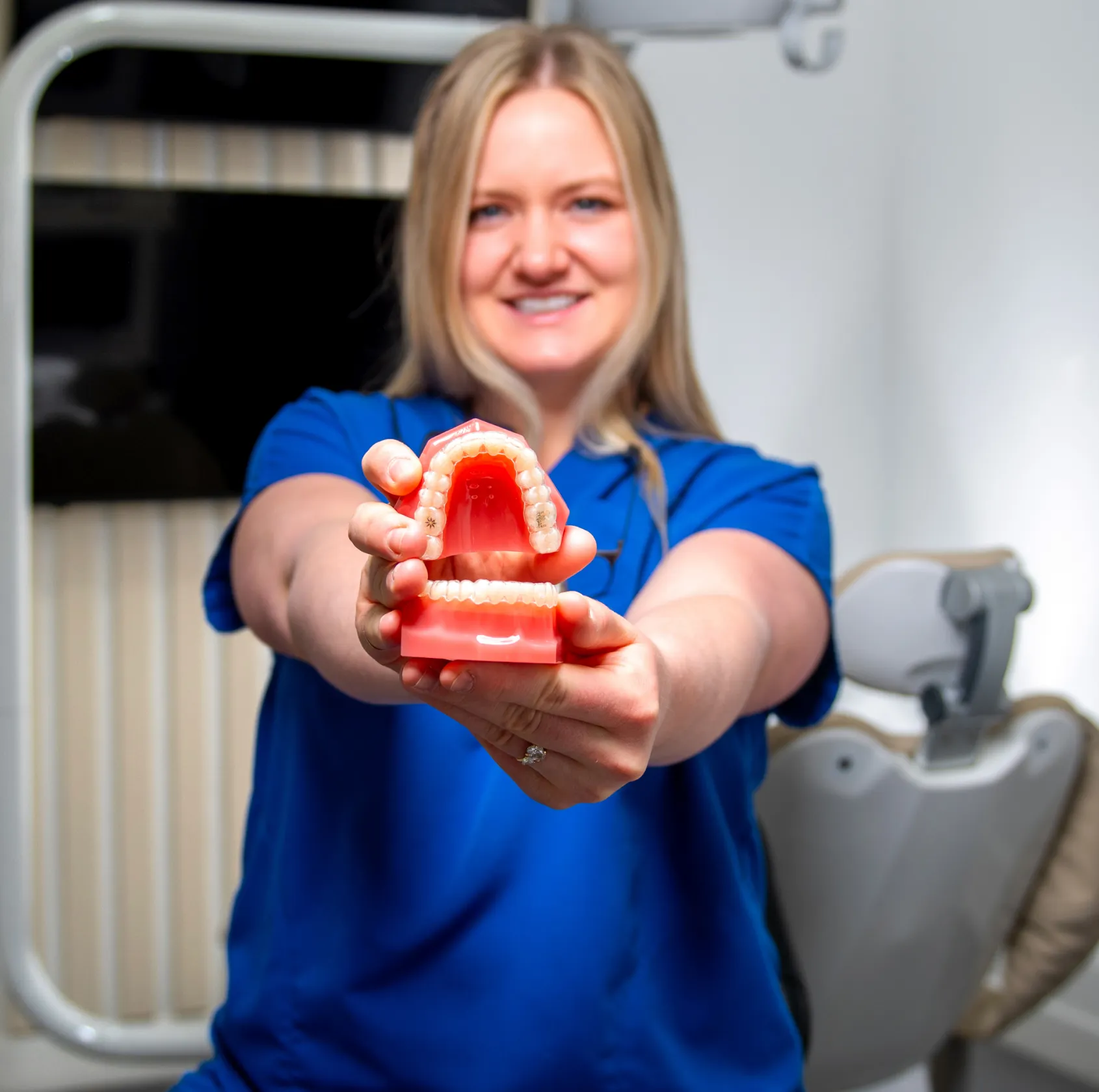 Smiling dental professional in blue scrubs holding a dental model with upper and lower teeth.