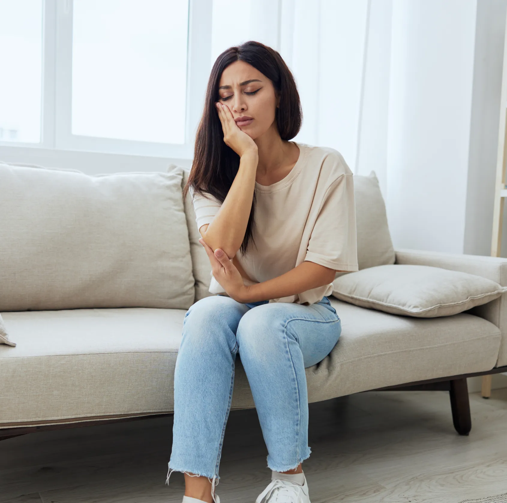 Woman sitting on a beige sofa holding her cheek in pain, appearing to have a toothache.