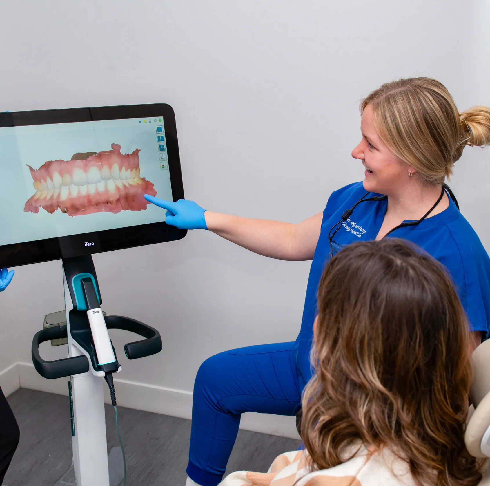 Dentist in blue scrubs pointing at 3D dental model on a screen while consulting a patient.