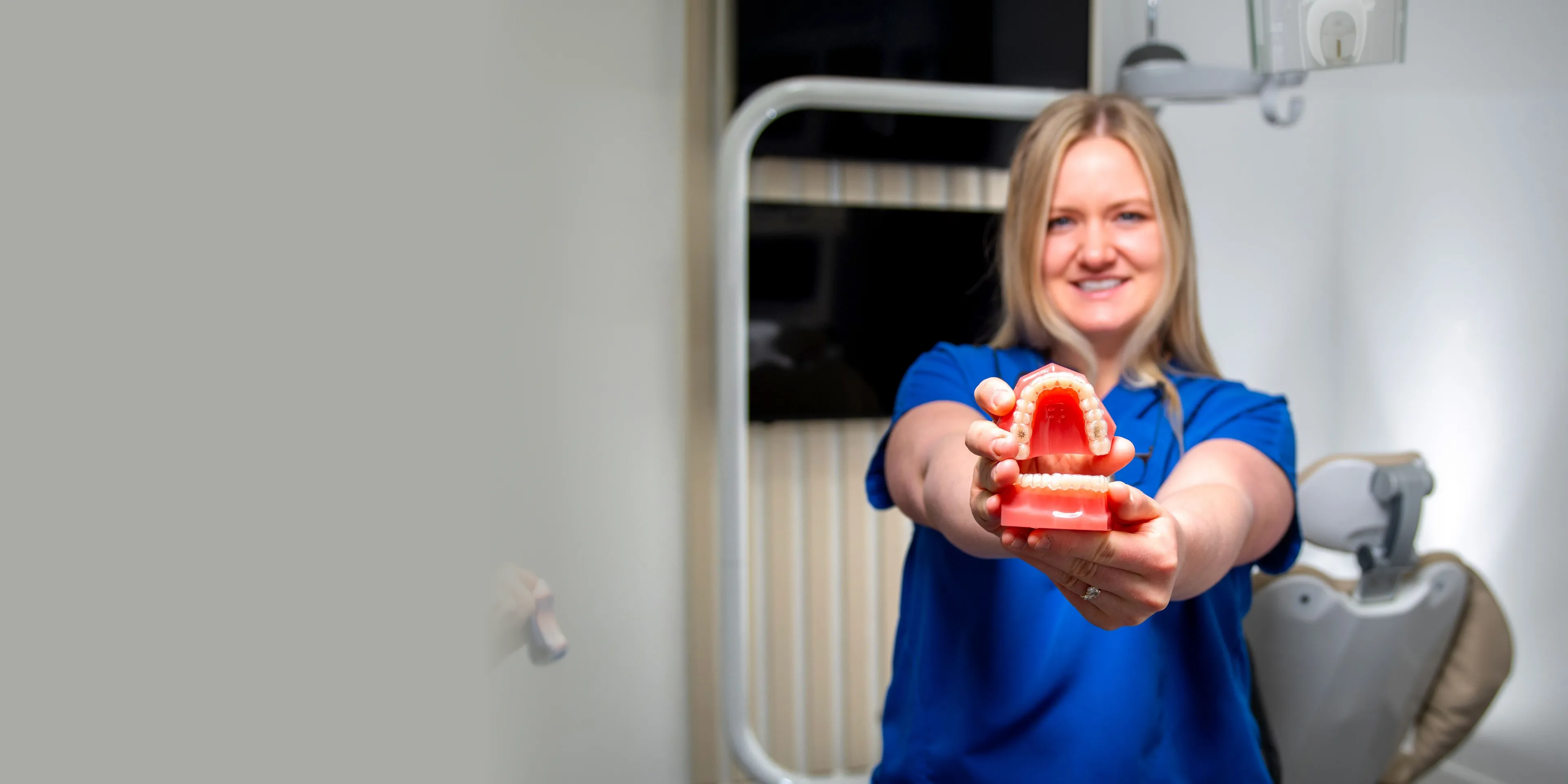 Smiling dental professional in blue scrubs holding out a dental model showing teeth and gums.