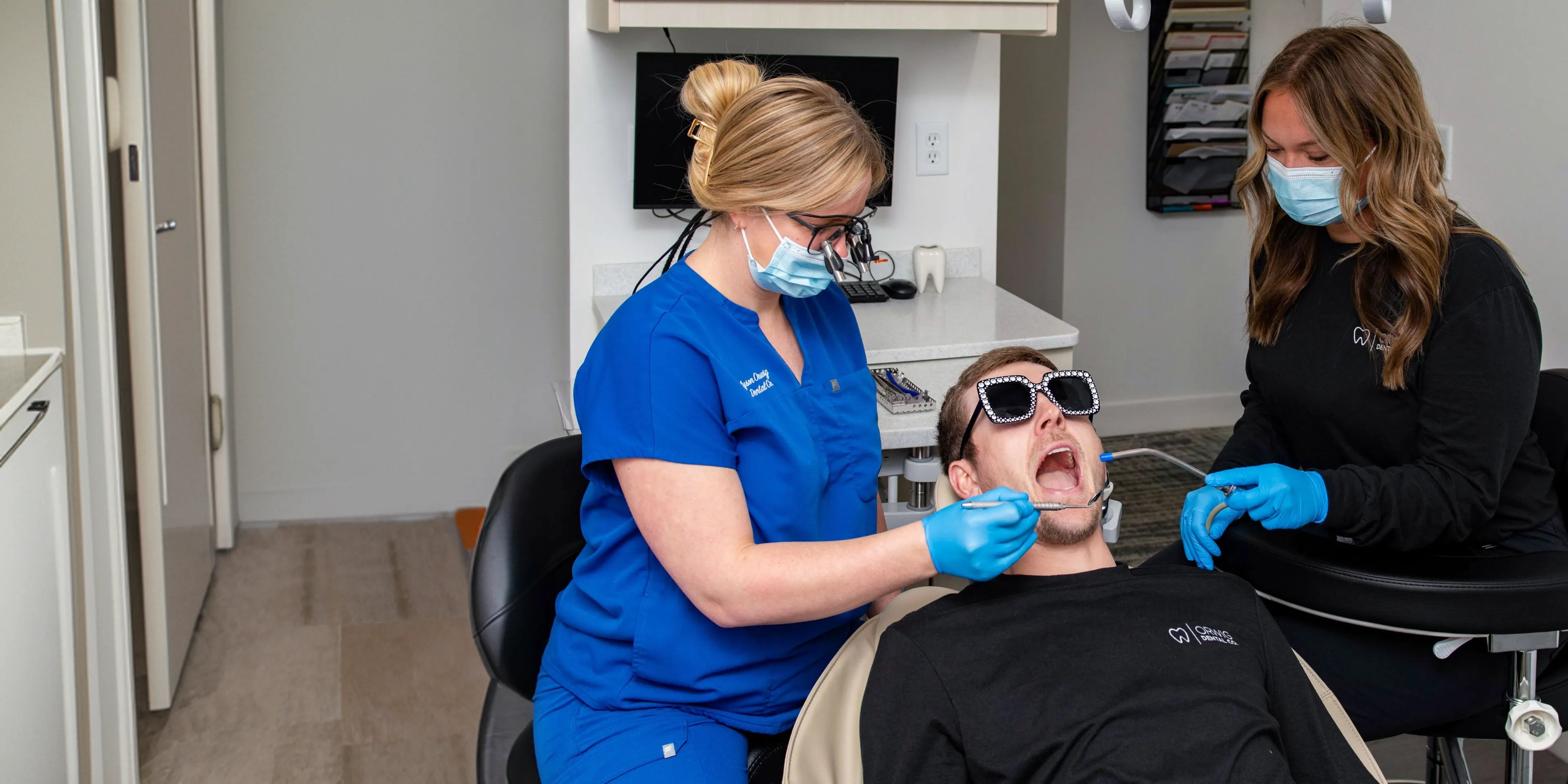 Dental hygienist in blue scrubs and mask cleaning a man's teeth while another assistant holds a suction tool during a dental checkup.