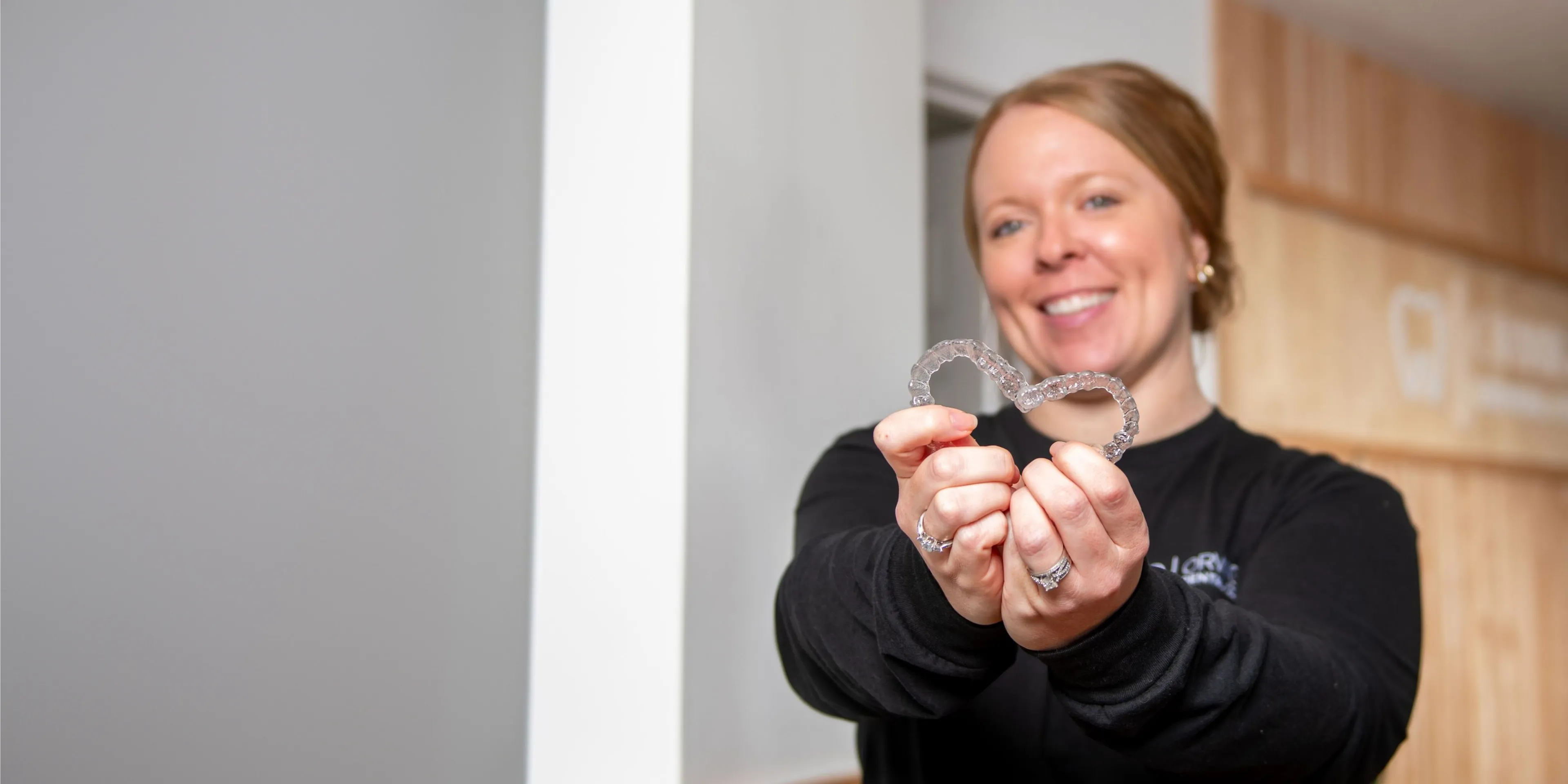 Smiling woman holding clear Invisalign aligners shaped like a heart towards the camera.